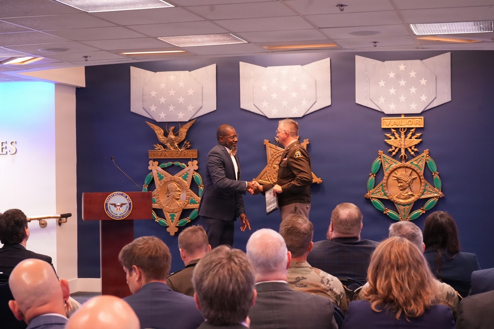 A formal ceremony is taking place in what appears to be a Department of Defense facility, featuring large Medal of Honor decorative displays on a blue wall. Two individuals — one in civilian attire and one in military uniform — are shaking hands on a stage near a podium bearing the Department of Defense seal. An audience of approximately 15-20 attendees, some in military uniforms and others in civilian dress, are seated facing the stage.
