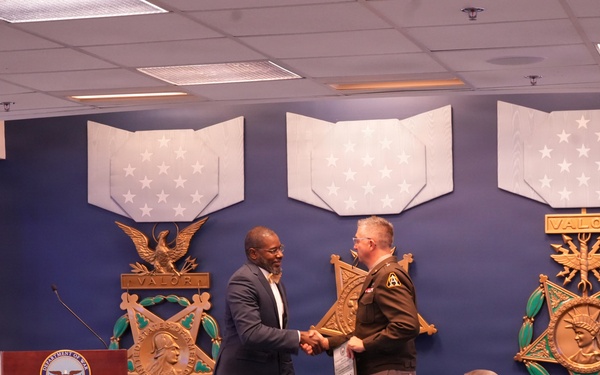 A formal ceremony is taking place in what appears to be a Department of Defense facility, featuring large Medal of Honor decorative displays on a blue wall. Two individuals — one in civilian attire and one in military uniform — are shaking hands on a stage near a podium bearing the Department of Defense seal. An audience of approximately 15-20 attendees, some in military uniforms and others in civilian dress, are seated facing the stage.