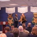 A formal ceremony is taking place in what appears to be a Department of Defense facility, featuring large Medal of Honor decorative displays on a blue wall. Two individuals — one in civilian attire and one in military uniform — are shaking hands on a stage near a podium bearing the Department of Defense seal. An audience of approximately 15-20 attendees, some in military uniforms and others in civilian dress, are seated facing the stage.