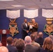 A formal military ceremony is taking place in what appears to be the Pentagon or a Department of Defense facility. Two individuals — one in civilian attire and one in military uniform — are shaking hands and holding a plaque or certificate together on stage. An audience of military and civilian personnel is seated in the foreground observing the event.