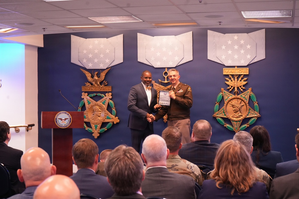 A formal award ceremony is taking place in what appears to be the Pentagon or a Department of Defense facility. Two individuals — one in civilian attire and one in U.S. Army uniform — are shaking hands and presenting a plaque or award on a stage. The backdrop features large decorative representations of the Medal of Honor for multiple military branches.