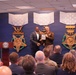 A formal award ceremony is taking place in what appears to be the Pentagon or a Department of Defense facility. Two individuals — one in civilian attire and one in U.S. Army uniform — are shaking hands and presenting a plaque or award on a stage. The backdrop features large decorative representations of the Medal of Honor for multiple military branches.
