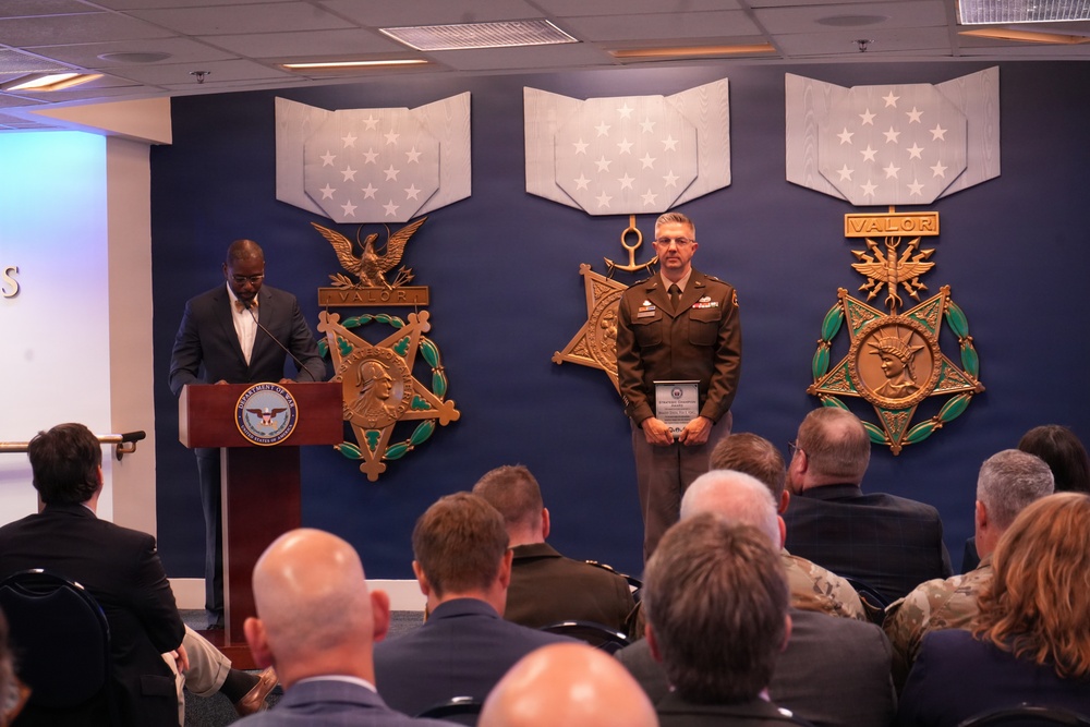 A formal military awards ceremony is taking place in what appears to be the Pentagon's Medal of Honor Hall or a similar official Department of Defense venue. A civilian official speaks at a podium bearing the Department of Defense seal while a military officer in Army service uniform stands holding an award plaque. An audience of military and civilian personnel is seated facing the stage.