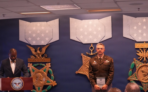 A formal military awards ceremony is taking place in what appears to be the Pentagon's Medal of Honor Hall or a similar official Department of Defense venue. A civilian official speaks at a podium bearing the Department of Defense seal while a military officer in Army service uniform stands holding an award plaque. An audience of military and civilian personnel is seated facing the stage.