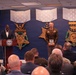 A formal military awards ceremony is taking place in what appears to be the Pentagon's Medal of Honor Hall or a similar official Department of Defense venue. A civilian official speaks at a podium bearing the Department of Defense seal while a military officer in Army service uniform stands holding an award plaque. An audience of military and civilian personnel is seated facing the stage.