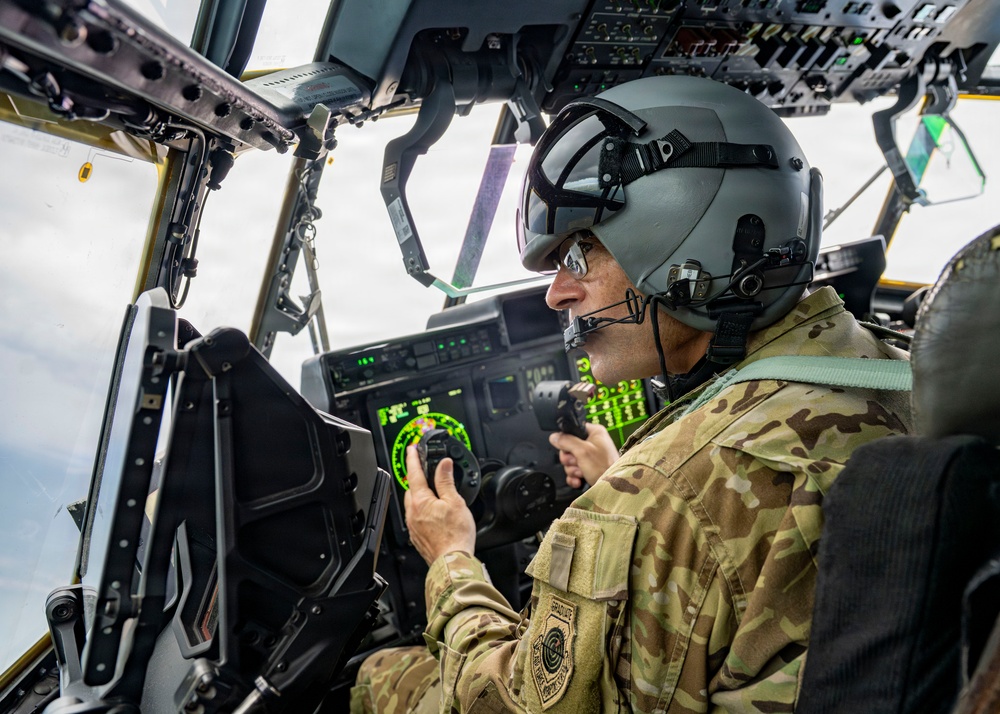 A military aviator is seated in the cockpit of a large military aircraft, wearing a flight helmet with integrated visor and communications equipment. The pilot is operating controls while monitoring multiple digital displays showing navigation and flight data. The cockpit features a complex array of instruments, screens, and overhead panels consistent with a large transport or special operations aircraft.