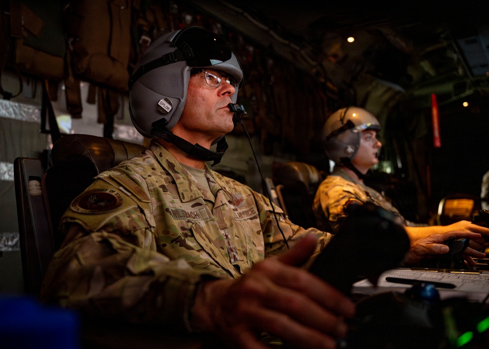 Two U.S. Air Force personnel are seated inside what appears to be a military aircraft, wearing flight helmets with communication equipment. The individual in the foreground has a name tape reading 'WILSBACH' and wears OCP (Operational Camouflage Pattern) uniform with Air Force patches. Both personnel appear to be monitoring or operating equipment at a workstation inside the aircraft.