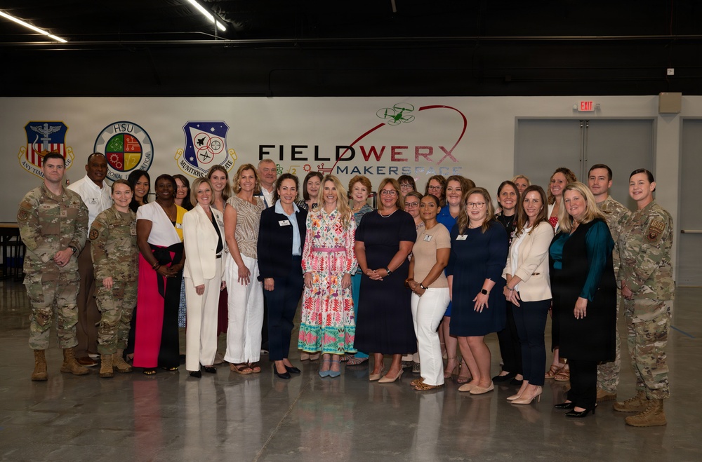 A large group of approximately 30 people pose for a formal group photograph at the FieldWerx Makerspace facility. The group includes both military personnel in camouflage uniforms and civilians in business/professional attire, suggesting an official visit or event. Multiple military unit insignia patches are visible on the wall behind them.