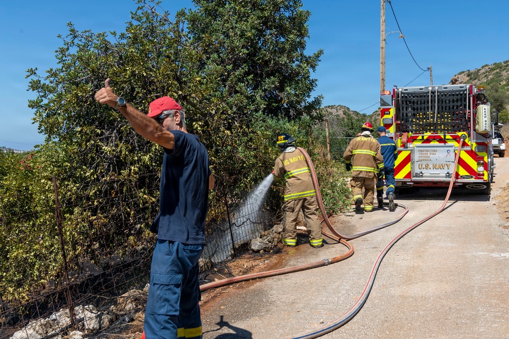 U.S. Navy firefighters are actively combating a vegetation fire along a roadside, using hoses connected to a U.S. Navy fire truck. A civilian or coordinator in a red cap and blue uniform is giving a thumbs-up gesture in the foreground, while two firefighters in full gear direct water at burning or smoldering brush near a wire fence. The scene appears to be in a Mediterranean or arid hillside environment under clear blue skies.