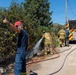 U.S. Navy firefighters are actively combating a vegetation fire along a roadside, using hoses connected to a U.S. Navy fire truck. A civilian or coordinator in a red cap and blue uniform is giving a thumbs-up gesture in the foreground, while two firefighters in full gear direct water at burning or smoldering brush near a wire fence. The scene appears to be in a Mediterranean or arid hillside environment under clear blue skies.