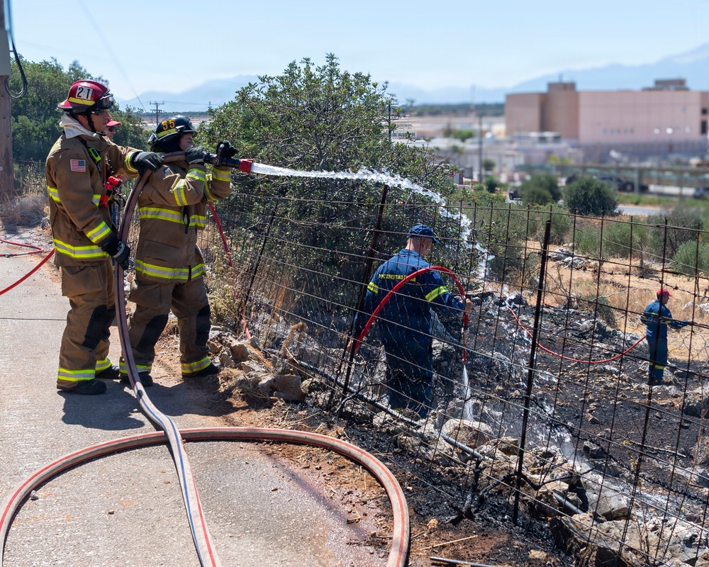 Firefighters are actively working to extinguish a brush or vegetation fire along a fenced area near a roadway. Two firefighters in full turnout gear are operating a hose nozzle from the road, while two additional personnel in blue uniforms work within the burned area beyond a wire fence. The scene shows significant char and ash on the ground, indicating the fire has been partially suppressed.