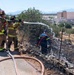 Firefighters are actively working to extinguish a brush or vegetation fire along a fenced area near a roadway. Two firefighters in full turnout gear are operating a hose nozzle from the road, while two additional personnel in blue uniforms work within the burned area beyond a wire fence. The scene shows significant char and ash on the ground, indicating the fire has been partially suppressed.