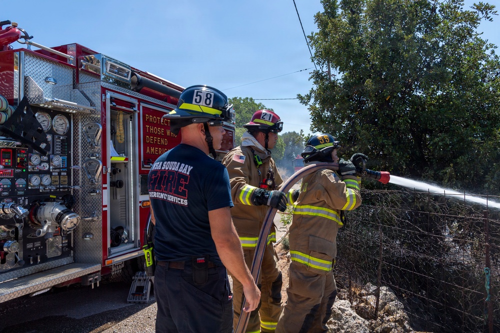 Three firefighters are actively responding to a fire near a fence line and dense vegetation. Two firefighters in full turnout gear are operating a hose line directing water toward the fire, while a third in a navy blue shirt and helmet monitors the operation. A red fire engine with visible pump panel gauges and the inscription 'Protect Those Who Defend America' is parked nearby.