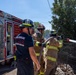 Three firefighters are actively responding to a fire near a fence line and dense vegetation. Two firefighters in full turnout gear are operating a hose line directing water toward the fire, while a third in a navy blue shirt and helmet monitors the operation. A red fire engine with visible pump panel gauges and the inscription 'Protect Those Who Defend America' is parked nearby.