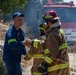 A firefighter in a blue uniform and cap is shaking hands with another firefighter wearing full turnout gear and a red helmet numbered '21'. A third firefighter wearing a helmet numbered '55' stands nearby. In the background, a red fire truck (Mercedes-Benz Atego) is visible amid smoke, suggesting an active or recently controlled fire incident.