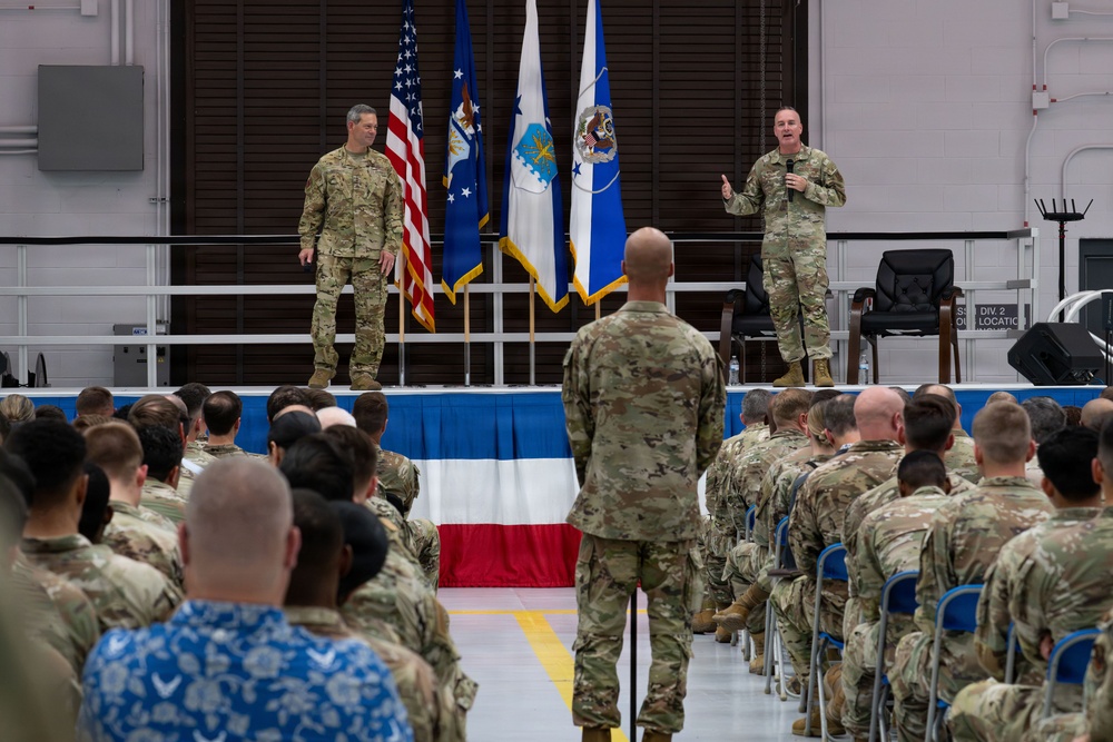 A military formation or all-hands meeting is taking place inside what appears to be an aircraft hangar or large military facility. Two uniformed personnel stand on a raised stage, with one speaking into a microphone while addressing a large seated audience of military members in camouflage uniforms. Multiple flags, including the U.S. flag and what appear to be Air Force flags, are displayed on the stage.