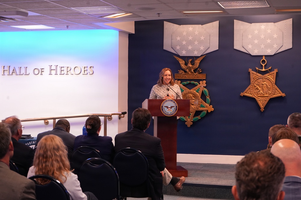 A woman is speaking at a podium bearing the Department of Defense seal in the Pentagon's Hall of Heroes. The backdrop features large decorative representations of the Medal of Honor — the Army version (left) and the Navy version (right) — mounted on a dark blue wall. An audience of attendees is seated facing the speaker during what appears to be an official ceremony or event.