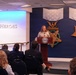 A woman is speaking at a podium bearing the Department of Defense seal in the Pentagon's Hall of Heroes. The backdrop features large decorative representations of the Medal of Honor — the Army version (left) and the Navy version (right) — mounted on a dark blue wall. An audience of attendees is seated facing the speaker during what appears to be an official ceremony or event.