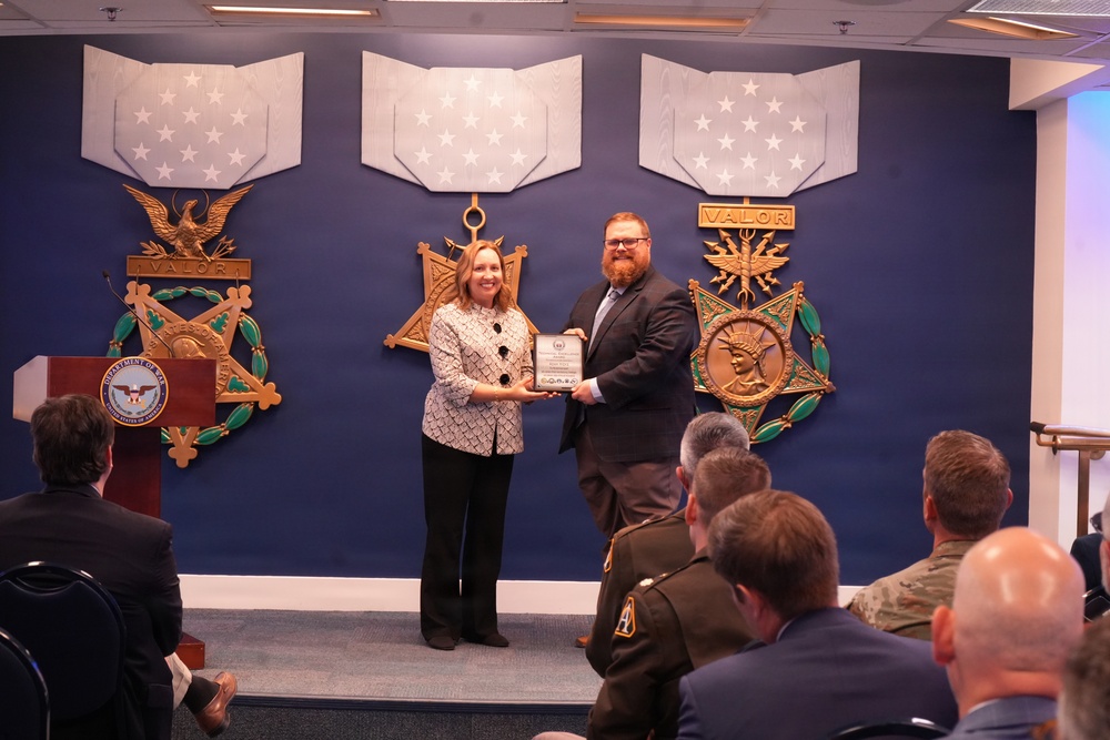 Two individuals stand on a stage holding a plaque or certificate during what appears to be an official award ceremony. The event is taking place in a formal government setting, likely the Pentagon, as evidenced by the Department of Defense seal on the podium and large Medal of Honor insignia displayed on the blue wall behind them. An audience that includes military personnel in uniform is seated in the foreground.