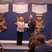 Two individuals stand on a stage holding a plaque or certificate during what appears to be an official award ceremony. The event is taking place in a formal government setting, likely the Pentagon, as evidenced by the Department of Defense seal on the podium and large Medal of Honor insignia displayed on the blue wall behind them. An audience that includes military personnel in uniform is seated in the foreground.