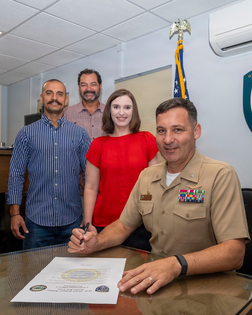 A U.S. Navy officer in khaki uniform is seated at a desk signing an official document, identified as a 'Commanding Officer's Proclamation' from what appears to be a Naval Support Activity. Three civilians — two men and one woman — stand behind him smiling, suggesting a formal ceremonial signing event. An American flag with gold fringe and a unit insignia shield are visible in the background.