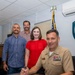 A U.S. Navy officer in khaki uniform is seated at a desk signing an official document, identified as a 'Commanding Officer's Proclamation' from what appears to be a Naval Support Activity. Three civilians — two men and one woman — stand behind him smiling, suggesting a formal ceremonial signing event. An American flag with gold fringe and a unit insignia shield are visible in the background.