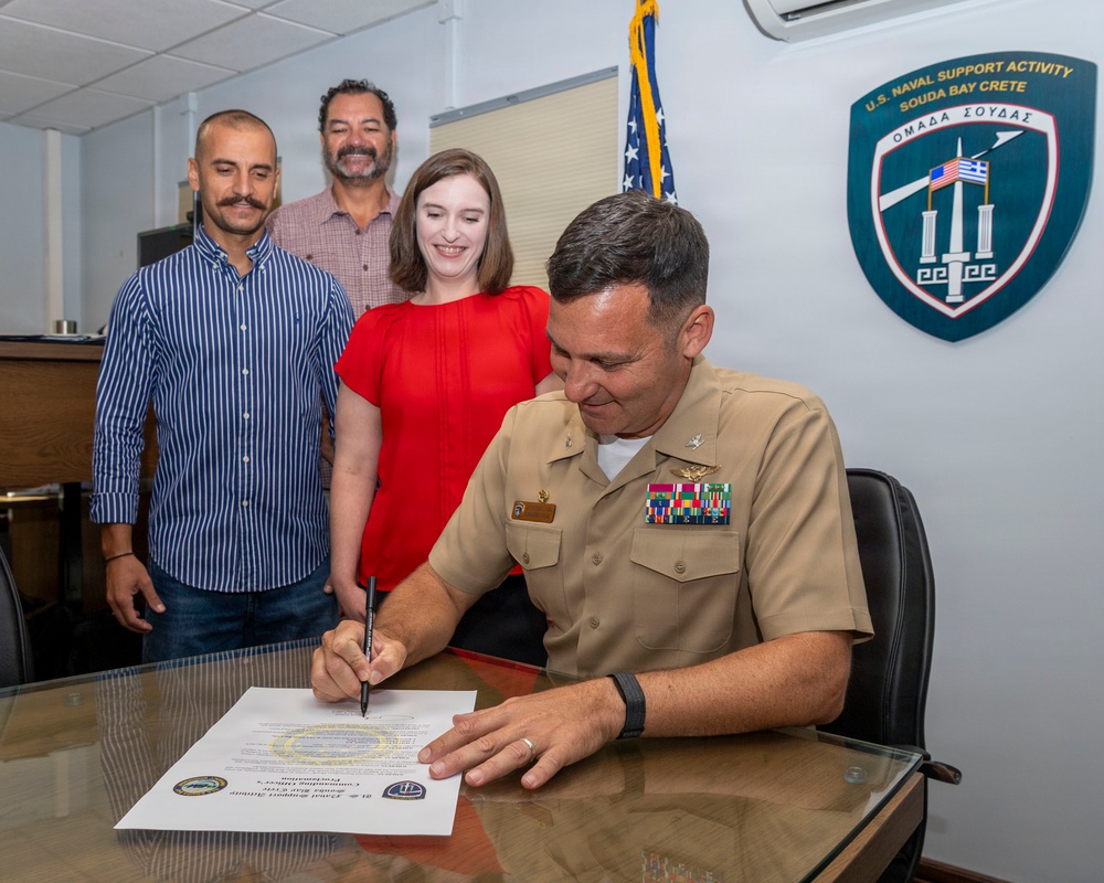 A U.S. Navy officer in khaki uniform is signing an official document at a desk, observed by three civilians standing behind him. The setting appears to be an office at U.S. Naval Support Activity Souda Bay, Crete, as indicated by the official emblem on the wall. The occasion appears to be a formal signing ceremony or proclamation.