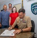 A U.S. Navy officer in khaki uniform is signing an official document at a desk, observed by three civilians standing behind him. The setting appears to be an office at U.S. Naval Support Activity Souda Bay, Crete, as indicated by the official emblem on the wall. The occasion appears to be a formal signing ceremony or proclamation.