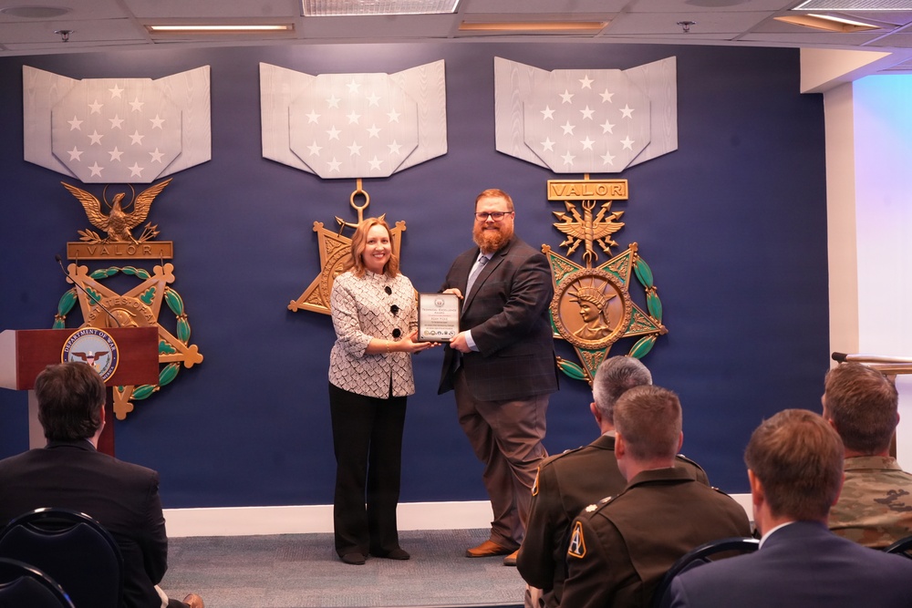 Two individuals are posing for a photo while jointly holding a framed award or certificate during what appears to be an official recognition ceremony. The event is taking place in a formal military or Department of Defense setting, evidenced by Medal of Honor display plaques on the wall behind them. An audience of military personnel and civilians is seated in the foreground.