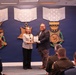 Two individuals are posing for a photo while jointly holding a framed award or certificate during what appears to be an official recognition ceremony. The event is taking place in a formal military or Department of Defense setting, evidenced by Medal of Honor display plaques on the wall behind them. An audience of military personnel and civilians is seated in the foreground.