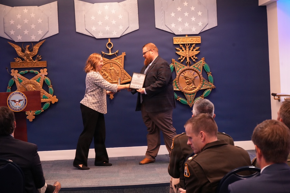 An award ceremony is taking place in what appears to be a Department of War or Department of Defense venue, with large decorative Medal of Honor replicas on the wall. A woman is presenting a framed certificate or plaque to a bearded man in a dark suit on a small stage. Several attendees, including individuals in military uniforms, are seated in the foreground observing the presentation.