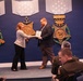 An award ceremony is taking place in what appears to be a Department of War or Department of Defense venue, with large decorative Medal of Honor replicas on the wall. A woman is presenting a framed certificate or plaque to a bearded man in a dark suit on a small stage. Several attendees, including individuals in military uniforms, are seated in the foreground observing the presentation.