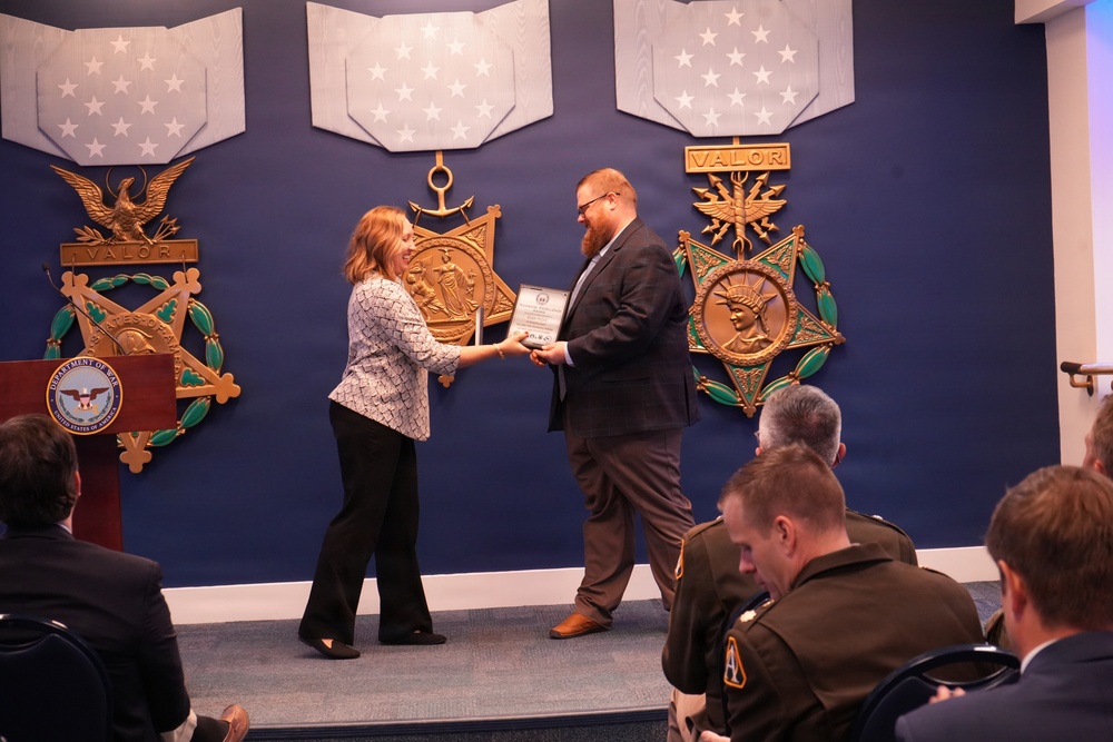 A formal award ceremony is taking place in what appears to be a Department of Defense or Pentagon venue. A woman in a patterned blazer is presenting a framed certificate or plaque to a bearded man in a dark suit. Military personnel and civilians are seated in the audience observing the presentation.