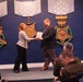 A formal award ceremony is taking place in what appears to be a Department of Defense or Pentagon venue. A woman in a patterned blazer is presenting a framed certificate or plaque to a bearded man in a dark suit. Military personnel and civilians are seated in the audience observing the presentation.