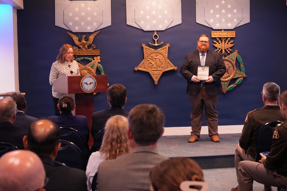 A formal award ceremony is taking place in what appears to be a Department of Defense facility, identifiable by the DoD seal on the podium and large Medal of Honor replica decorations on the blue wall. A woman is speaking at the podium while a bearded man in a suit holds a framed award or plaque. An audience of attendees, including at least one uniformed military officer, is seated facing the stage.
