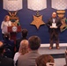 A formal award ceremony is taking place in what appears to be a Department of Defense facility, identifiable by the DoD seal on the podium and large Medal of Honor replica decorations on the blue wall. A woman is speaking at the podium while a bearded man in a suit holds a framed award or plaque. An audience of attendees, including at least one uniformed military officer, is seated facing the stage.