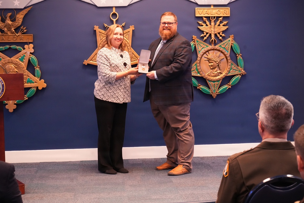Two civilians stand before a blue wall decorated with large gold Medal of Honor emblems and 'VALOR' inscriptions, jointly holding an open medal presentation case. The setting appears to be a formal military awards ceremony, likely at the Pentagon or a similar Department of Defense facility. An audience including at least one uniformed military officer is visible in the foreground.