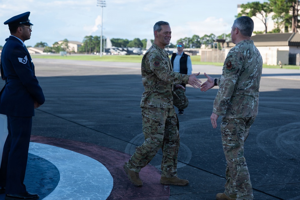 Two military personnel in OCP (Operational Camouflage Pattern) uniforms are greeting each other with a handshake on an airfield tarmac. A third service member in dress blues stands at attention to the left, while a civilian in dark clothing observes in the background. The setting appears to be a formal arrival or greeting ceremony at a military air base.
