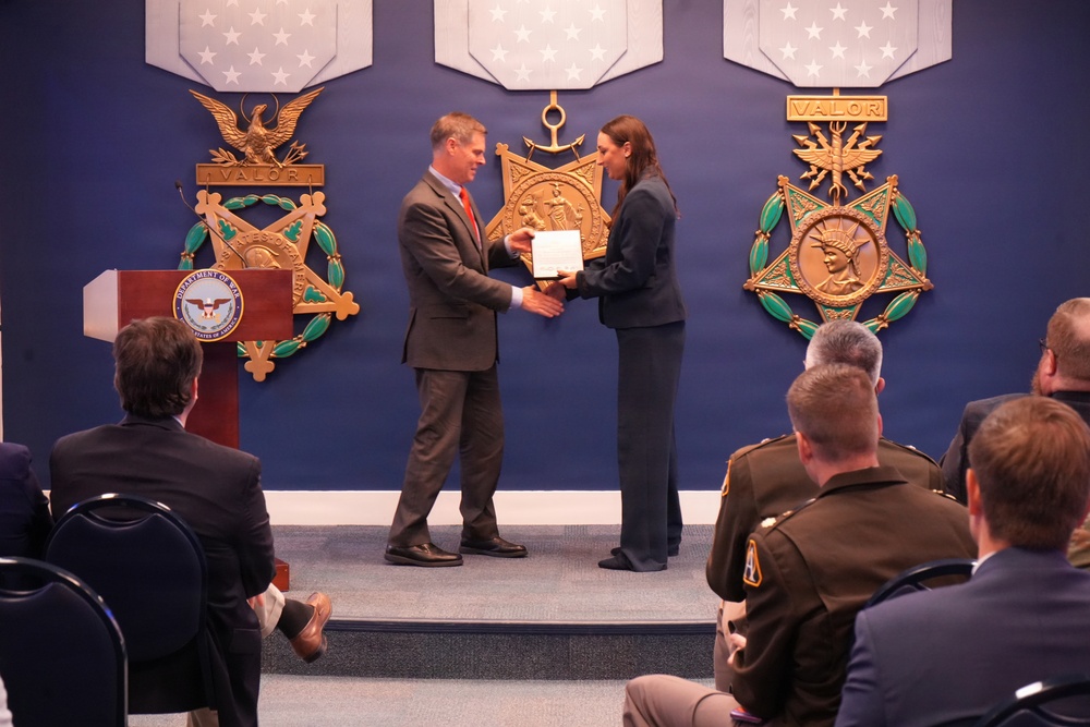 A formal ceremony is taking place at what appears to be the Pentagon or a Department of War/Defense facility, where a man in a gray suit with a red tie is presenting a document or certificate to a woman in a dark blazer. The backdrop features large decorative Medal of Honor emblems with 'VALOR' inscriptions and a Department of War seal on the podium. An audience of military personnel and civilians is seated watching the presentation.