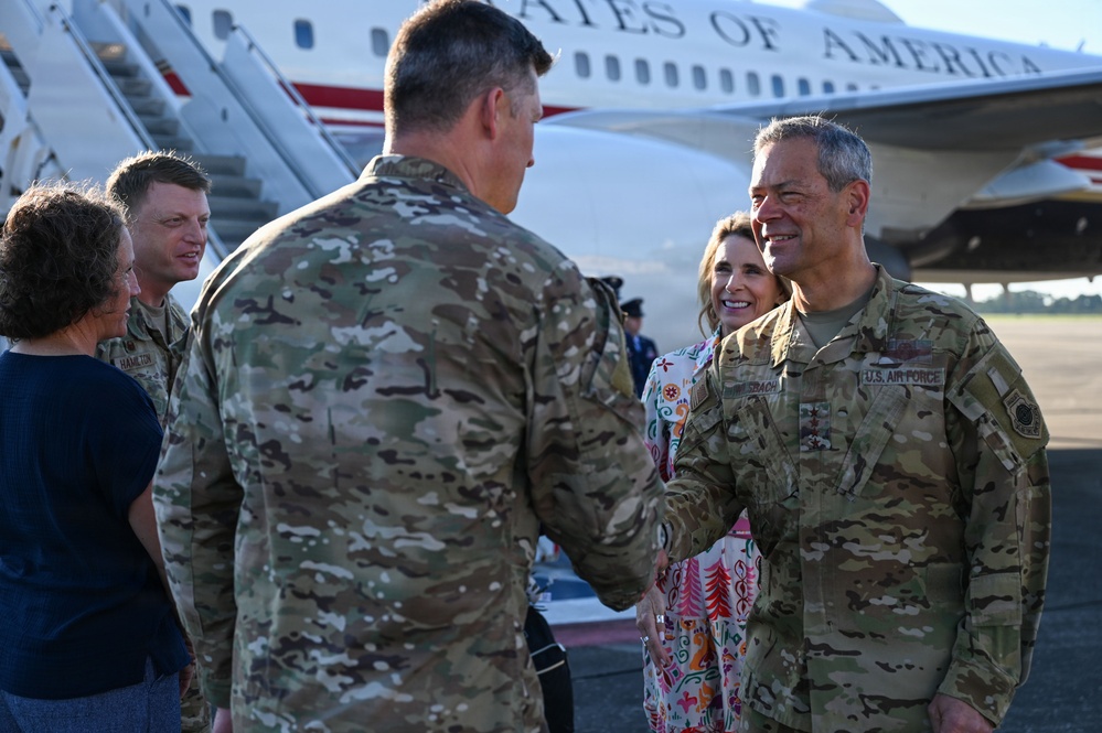 A group of U.S. Air Force personnel in OCP (Operational Camouflage Pattern) uniforms are gathered on an airport tarmac, engaged in a greeting or welcoming ceremony. A U.S. government aircraft bearing the text 'STATES OF AMERICA' is visible in the background with boarding stairs deployed. The scene appears to be an official arrival or departure event involving senior military officers.