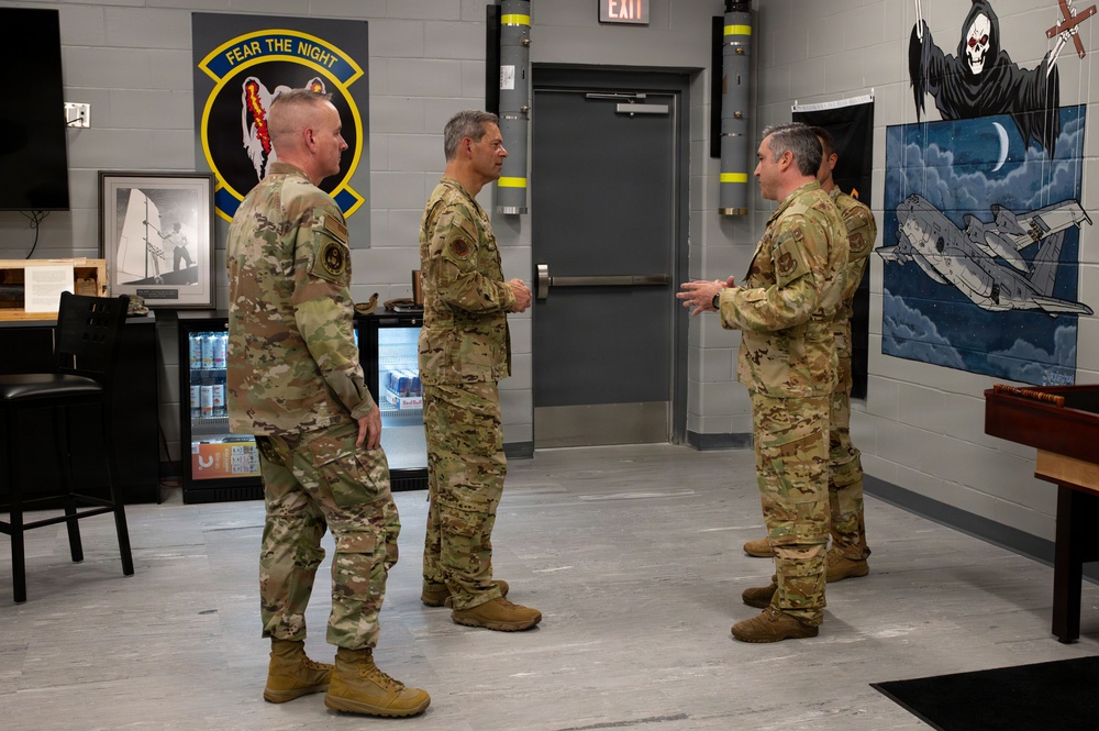Three military personnel in OCP (Operational Camouflage Pattern) uniforms are engaged in conversation inside what appears to be a squadron ready room or lounge area. The room features a unit patch with the motto 'Fear The Night' on the wall, along with military artwork depicting aircraft and a grim reaper motif. The setting suggests an informal briefing or meet-and-greet within a military aviation unit facility.