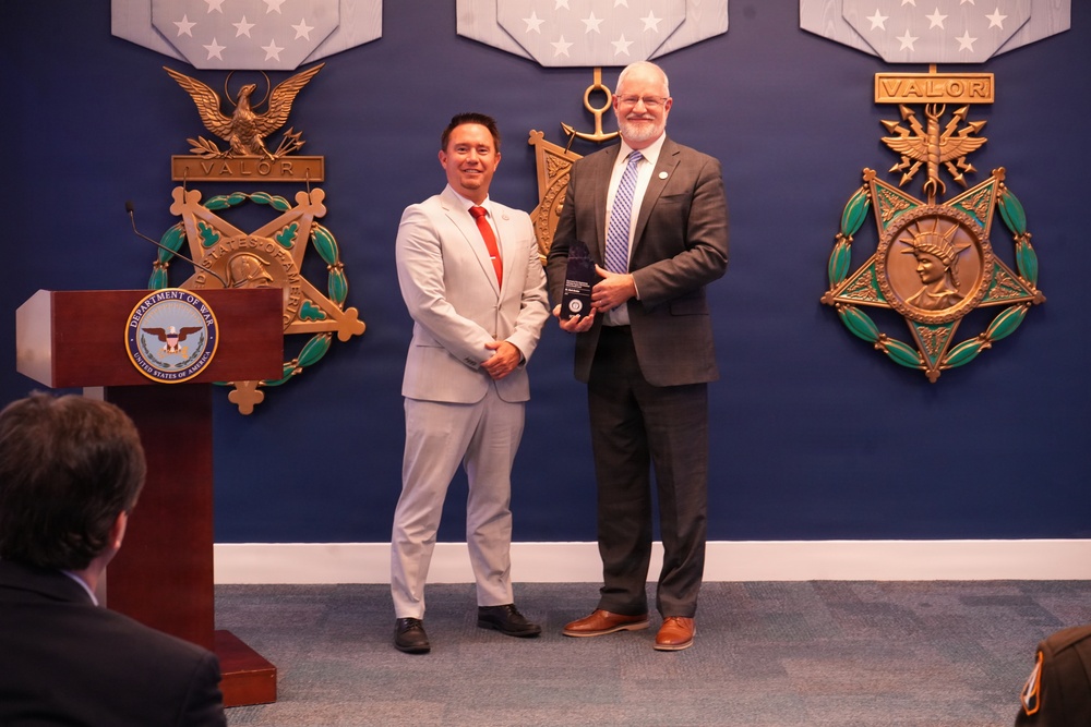 Two men are posing for a photo at what appears to be an official Department of Veterans Affairs award ceremony. One man in a light gray suit and the other in a dark gray suit are jointly holding a plaque or award. The backdrop features large decorative Medal of Honor replicas and a Department of Veterans Affairs podium.