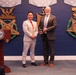 Two men are posing for a photo at what appears to be an official Department of Veterans Affairs award ceremony. One man in a light gray suit and the other in a dark gray suit are jointly holding a plaque or award. The backdrop features large decorative Medal of Honor replicas and a Department of Veterans Affairs podium.