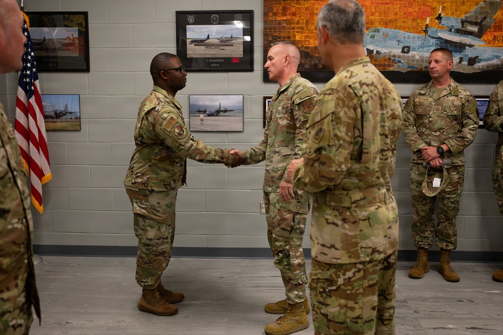 A formal military ceremony is taking place indoors at what appears to be a U.S. Air Force facility. Two service members in OCP (Operational Camouflage Pattern) uniforms are shaking hands in what appears to be a recognition or greeting ceremony. Several other uniformed personnel are observing in the background, with framed aviation photographs and a U.S. flag visible on the walls.