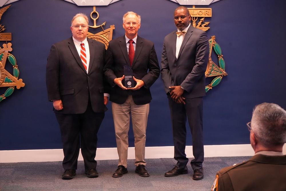 Three men in business attire are posing for a formal photograph in what appears to be a government or military building. The central figure is holding a dark award or plaque, suggesting this is an award ceremony or recognition event. A fourth individual, partially visible in the foreground, appears to be wearing a military uniform.