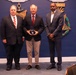 Three men in business attire are posing for a formal photograph in what appears to be a government or military building. The central figure is holding a dark award or plaque, suggesting this is an award ceremony or recognition event. A fourth individual, partially visible in the foreground, appears to be wearing a military uniform.