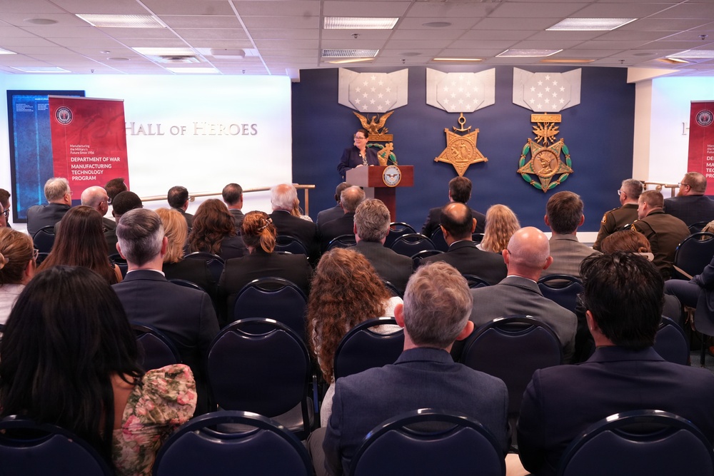 A formal presentation or ceremony is taking place in the Pentagon's Hall of Heroes, as indicated by the text on the blue wall backdrop. A speaker stands at a podium addressing a large audience seated in rows of chairs. The event appears to be related to the Department of War Manufacturing Technology Program, based on visible signage.