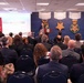 A formal presentation or ceremony is taking place in the Pentagon's Hall of Heroes, as indicated by the text on the blue wall backdrop. A speaker stands at a podium addressing a large audience seated in rows of chairs. The event appears to be related to the Department of War Manufacturing Technology Program, based on visible signage.