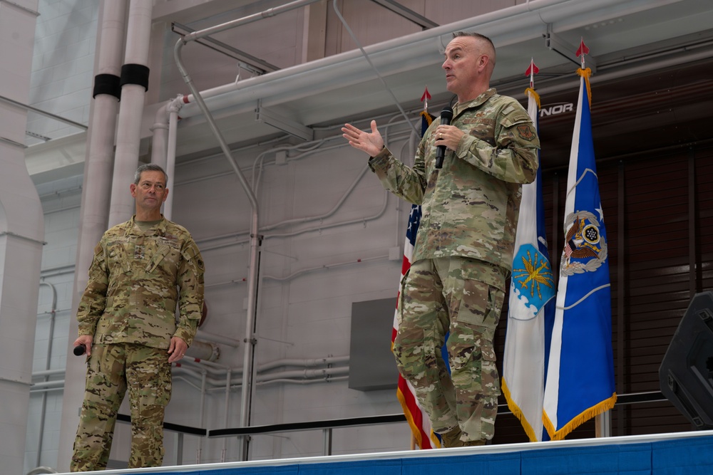Two military personnel in OCP (Operational Camouflage Pattern) uniforms are standing on a raised platform or stage in what appears to be a hangar or large indoor military facility. One individual is speaking into a microphone with gesturing hands, while the other stands at attention holding a microphone at his side. Several flags, including the American flag and what appears to be a U.S. Air Force flag, are visible in the background.