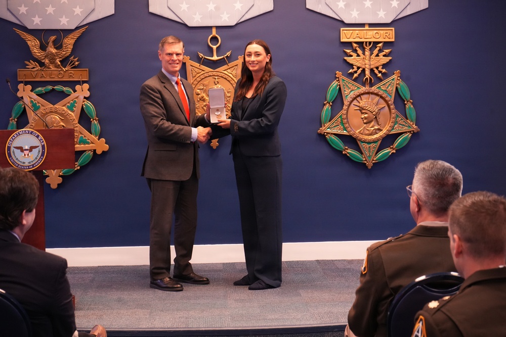 A formal award ceremony is taking place in what appears to be a U.S. Department of Defense facility. A man in a gray suit with an orange tie is presenting a medal or award in a display box to a woman in a dark suit, both shaking hands. Military personnel and civilians are seated in the audience observing the ceremony.